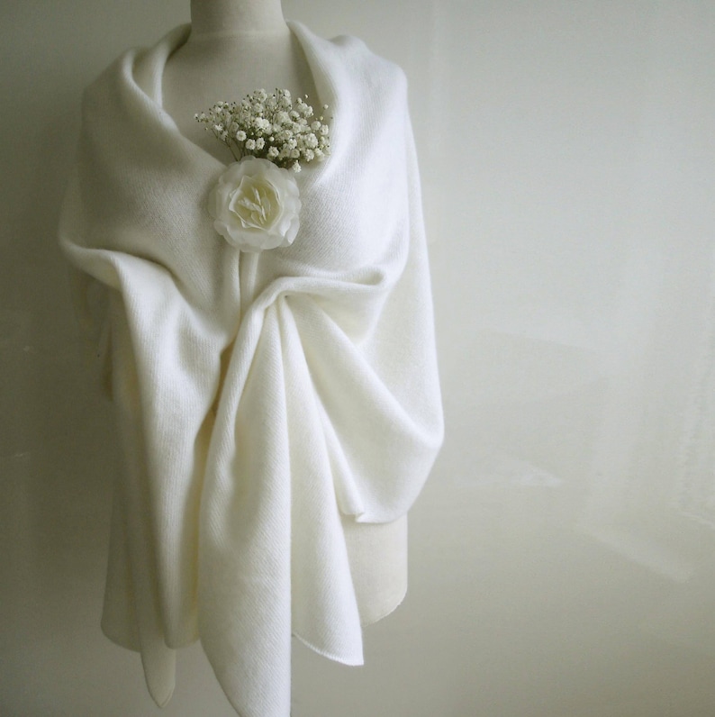 Puede incluir: Un chal color crema drapeado sobre un maniqu&iacute;, adornado con una rosa blanca y peque&ntilde;as flores blancas. El chal est&aacute; hecho de una tela suave y texturizada, creando pliegues elegantes. La est&eacute;tica general es delicada y refinada.