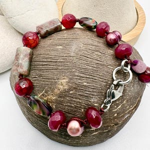 Pode incluir: Uma pulseira de contas com uma mistura de contas vermelhas, rosa e verdes. A pulseira inclui contas redondas, retangulares e ovais, com um fecho prateado. As contas s&atilde;o enfiadas num cord&atilde;o castanho escuro.