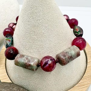 Pode incluir: Uma pulseira de contas com contas bord&ocirc;, verdes e rosas. A pulseira inclui contas grandes, redondas e retangulares. As contas s&atilde;o enfiadas com um cord&atilde;o escuro. A pulseira &eacute; exibida em um expositor de joias bege.