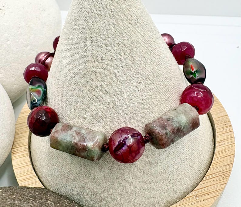 Pode incluir: Uma pulseira de contas com contas bord&ocirc;, verdes e rosas. A pulseira inclui contas grandes, redondas e retangulares. As contas s&atilde;o enfiadas com um cord&atilde;o escuro. A pulseira &eacute; exibida em um expositor de joias bege.