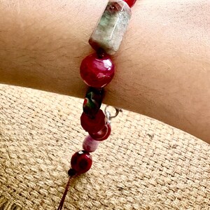 Pode incluir: Uma pulseira de contas com uma mistura de contas vermelhas, rosa e verdes. A pulseira inclui contas grandes, redondas e retangulares. As contas s&atilde;o enfiadas com uma corrente prateada e um fio castanho.