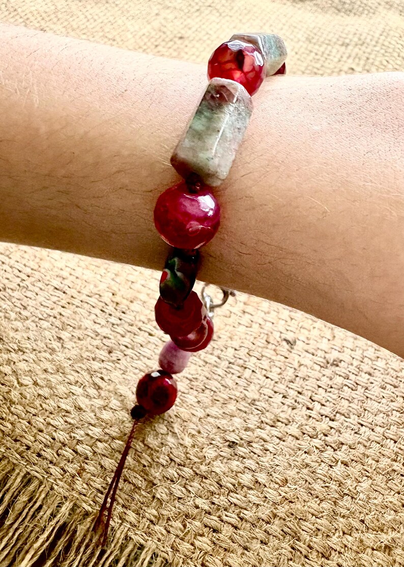 Pode incluir: Uma pulseira de contas com uma mistura de contas vermelhas, rosa e verdes. A pulseira inclui contas grandes, redondas e retangulares. As contas s&atilde;o enfiadas com uma corrente prateada e um fio castanho.