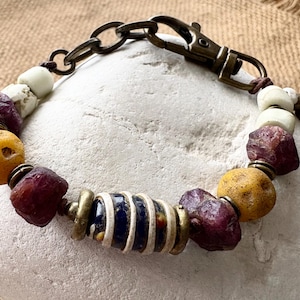 May include: A bracelet featuring a mix of raw, deep red ruby-like stones, smooth yellow spherical beads, white cylindrical beads, and a central dark blue barrel-shaped bead with white stripes, all connected by antique brass metal spacers and a clasp.