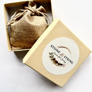 Pode incluir: Uma caixa de joias bege com a tampa aberta, revelando um pequeno saco de juta no interior. A caixa tem um adesivo circular com o texto "STONE & STRING Handcrafted Jewelry". O saco &eacute; amarrado com um cord&atilde;o. A caixa est&aacute; sobre uma superf&iacute;cie branca.