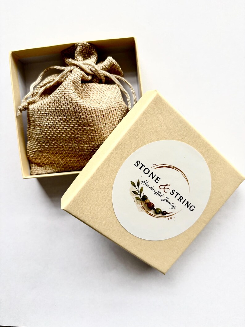 Pode incluir: Uma caixa de joias bege com a tampa aberta, revelando um pequeno saco de juta no interior. A caixa tem um adesivo circular com o texto "STONE & STRING Handcrafted Jewelry". O saco &eacute; amarrado com um cord&atilde;o. A caixa est&aacute; sobre uma superf&iacute;cie branca.