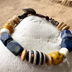 May include: A bracelet featuring a mix of blue lapis lazuli, yellow, and white beads strung on a brown cord. The design includes a central striped bead, metallic spacers, and a circular metal button closure, creating a textured appearance.