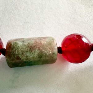Pode incluir: Close-up de um colar com uma conta cil&iacute;ndrica verde e duas contas redondas vermelhas. As contas s&atilde;o enfiadas em um cord&atilde;o marrom escuro, criando um contraste com o fundo branco. A conta verde tem uma apar&ecirc;ncia manchada.
