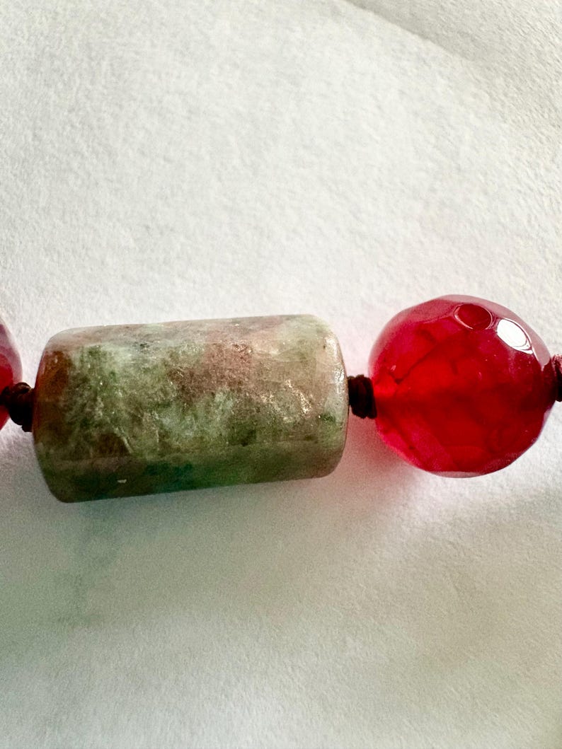 Pode incluir: Close-up de um colar com uma conta cil&iacute;ndrica verde e duas contas redondas vermelhas. As contas s&atilde;o enfiadas em um cord&atilde;o marrom escuro, criando um contraste com o fundo branco. A conta verde tem uma apar&ecirc;ncia manchada.