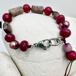 May include: A beaded bracelet featuring a variety of burgundy and pink beads, including faceted, round, and oblong shapes. The bracelet has a silver-tone clasp and is strung on a dark brown cord. Some beads have a pearlescent sheen.
