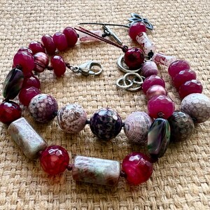 Pode incluir: Colar de contas com uma variedade de contas de pedra natural em tons de vermelho, roxo e cinza. O colar inclui contas redondas, ovais e retangulares, com um fecho prateado e detalhes decorativos. As contas s&atilde;o de v&aacute;rios tamanhos.