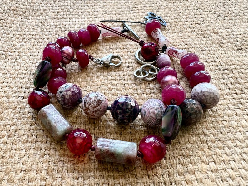 Pode incluir: Colar de contas com uma variedade de contas de pedra natural em tons de vermelho, roxo e cinza. O colar inclui contas redondas, ovais e retangulares, com um fecho prateado e detalhes decorativos. As contas s&atilde;o de v&aacute;rios tamanhos.