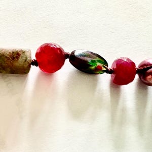 Pode incluir: Um close-up de um colar de contas com uma variedade de contas coloridas. O colar inclui uma conta retangular, salpicada de verde e marrom, contas vermelhas facetadas vibrantes e uma conta oval escura e iridescente. As contas s&atilde;o enfiadas em um cord&atilde;o escuro.