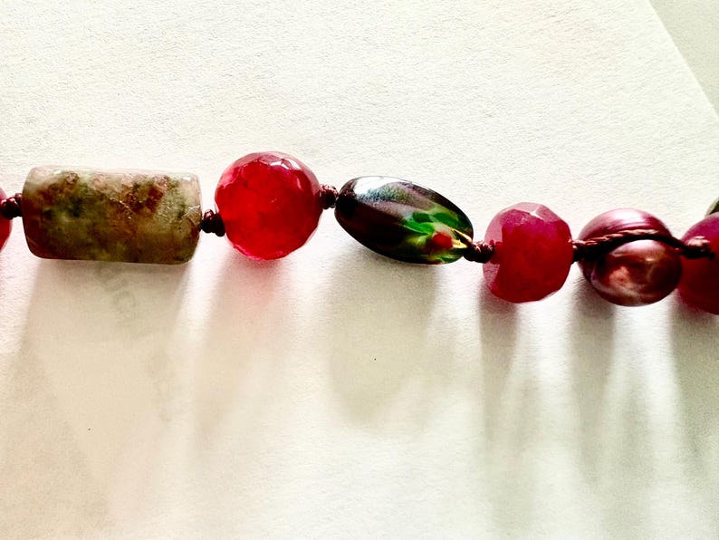 Pode incluir: Um close-up de um colar de contas com uma variedade de contas coloridas. O colar inclui uma conta retangular, salpicada de verde e marrom, contas vermelhas facetadas vibrantes e uma conta oval escura e iridescente. As contas s&atilde;o enfiadas em um cord&atilde;o escuro.