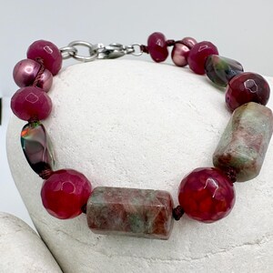 Pode incluir: Uma pulseira de contas com uma mistura de pedras vermelhas, rosa e verdes. A pulseira inclui contas vermelhas facetadas, pedras retangulares verdes e vermelhas e contas rosas semelhantes a p&eacute;rolas. &Eacute; enfiada em um cord&atilde;o marrom escuro e tem um fecho prateado.
