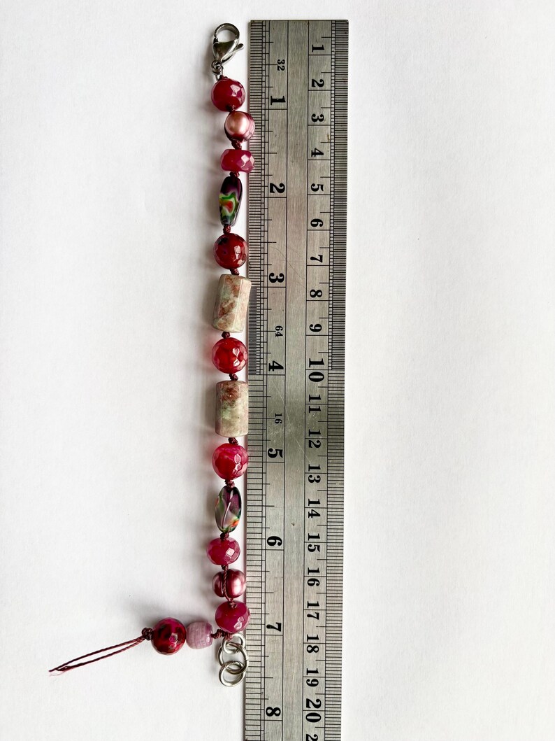 Pode incluir: Uma pulseira de contas com contas vermelhas, rosa e estampadas. A pulseira tem um fecho de lagosta e mede aproximadamente 18 cm de comprimento. As contas s&atilde;o de v&aacute;rias formas e tamanhos, com uma mistura de texturas.