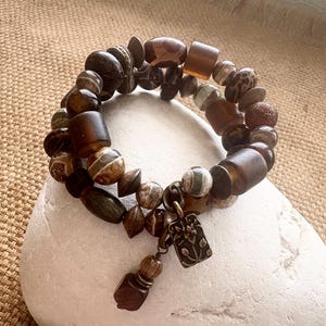 May include: A multi-strand bracelet featuring an assortment of brown, tan, and grey beads in various shapes including round, cylindrical, and faceted styles. A rectangular bronze-toned metal charm with an embossed floral design hangs from the bracelet.