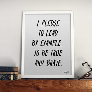 Peut inclure: Une impression encadrée blanche avec le texte "I pledge to lead by example, to be true and brave." sur un fond gris. L'impression est sur une étagère en bois avec un petit pot en céramique blanche avec une plante verte et un objet métallique.