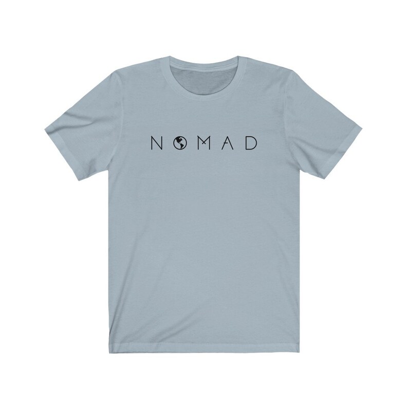 Puede incluir: Una camiseta azul claro con la palabra "NOMAD" impresa en letras negras. La "O" de "NOMAD" est&aacute; reemplazada por un peque&ntilde;o gr&aacute;fico de globo terr&aacute;queo negro.