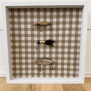 May include: A square white shadow box frame containing three vintage fishing lures mounted on a tan and white gingham fabric background. The top lure is metallic gold, the middle lure features black feathers, and the bottom lure is silver.