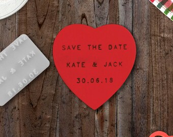 Pode incluir: Cartão de guardar a data em forma de coração vermelho com o texto "Save the Date Kate & Jack 30.06.18" sobre um fundo de madeira.