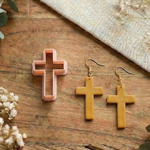 Puede incluir: Un cortador de galletas en forma de cruz rosa y un par de pendientes de cruz dorados. Los artículos están dispuestos sobre una superficie de madera con detalles florales.