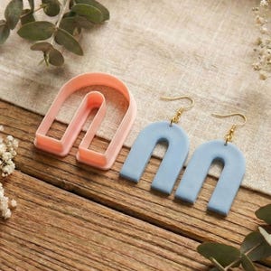 May include: Pale blue earrings with gold-coloured hooks, alongside a pink U-shaped clay cutter. The earrings and cutter are on a wooden surface, with sprigs of greenery and white flowers.
