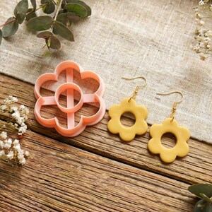May include: A flower-shaped cookie cutter and a pair of daisy earrings are displayed on a wooden surface. The cookie cutter is light pink, and the earrings are yellow with gold-colored hooks. Eucalyptus and small white flowers are arranged around the items.