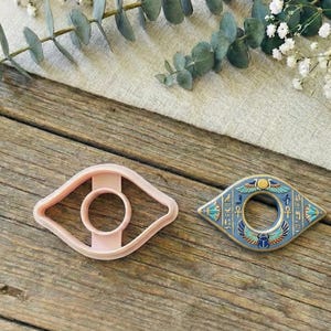 Op de afbeelding: Twee oogvormige objecten liggen op een houten oppervlak. Eén is een lichtroze koekjesvorm, de andere is een decoratief metalen stuk met blauwe, gouden en blauwgroene accenten. Eucalyptus en witte bloemen zijn op de achtergrond.