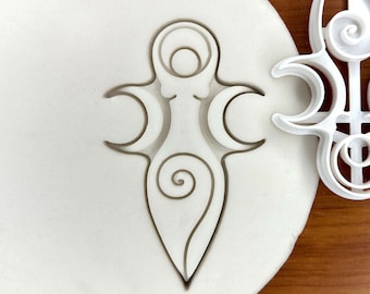 Puede incluir: Cortador de galletas de plástico blanco en forma de un símbolo de triple luna estilizado con un diseño en espiral. El cortador se muestra junto a una estera de horneado de silicona con el mismo diseño.