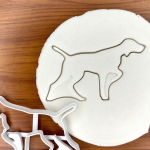 May include: White plastic cookie cutter in the shape of a German Shorthaired Pointer dog. The cutter is shown on a white surface with a silhouette of the dog.