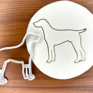 May include: Two white plastic cookie cutters shaped like dogs. The larger cutter is of a dog standing with its legs straight. The smaller cutter is of a dog sitting with its legs bent.