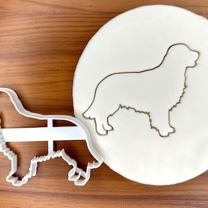 May include: White plastic cookie cutter in the shape of a golden retriever dog. The cutter is on a wooden surface next to a white fondant sheet with a gray outline of the same dog shape.