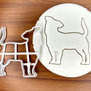 Puede incluir: Dos cortadores de galletas de plástico blanco en forma de perro. El cortador de la izquierda tiene un contorno detallado del perro, mientras que el cortador de la derecha es una silueta sólida.