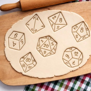Puede incluir: Un rodillo de madera descansa sobre una tabla de cortar de madera con una lámina de masa. La masa tiene varias formas de dados presionadas, incluyendo un dado de cuatro caras, un dado de seis caras y un dado de veinte caras. La masa es beige.