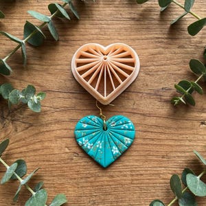 Puede incluir: Un pendiente turquesa en forma de corazón con un gancho dorado y un cortador de galletas a juego en forma de corazón. El pendiente tiene un estampado floral y está rodeado de hojas de eucalipto sobre una superficie de madera.