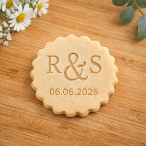 Può includere: Un biscotto rotondo con bordo smerlato di colore beige chiaro. Il centro presenta le lettere R & S in rilievo con la data 06.06.2026 stampata appena sotto le lettere in un carattere chiaro e senza grazie, creando un design personalizzato.