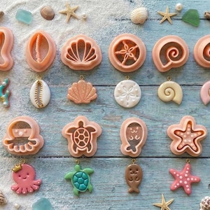 Puede incluir: Una colección de cortadores de galletas rosas con forma de concha, junto con estrellas de mar, conchas y vidrio marino, dispuestos sobre una superficie arenosa y un fondo de madera azul. Los cortadores incluyen varios diseños de criaturas marinas y conchas.