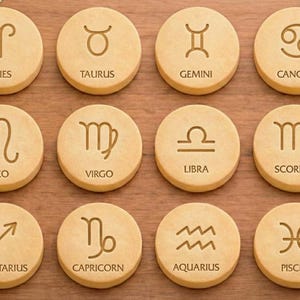 May include: Twelve round wooden coasters, each engraved with a different zodiac symbol and its name. The symbols are in a dark brown color, set against a light brown background. The coasters are arranged in three rows of four.