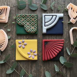 May include: A selection of cookie cutters and decorated biscuits. The cutters are shaped like a fan, a heart, and architectural designs. The biscuits are square with various patterns and colours, including green, yellow, red, and blue. Eucalyptus leaves are scattered around the items.