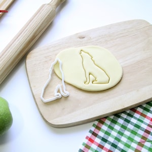 Puede incluir: Un cortador de galletas de lobo de plástico blanco sobre una tabla de cortar de madera con una pieza de masa extendida en forma de lobo. Hay un rodillo y una manzana verde en la encimera.