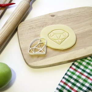 May include: A white plastic diamond-shaped cookie cutter on a wooden chopping board with a rolled out piece of dough in the shape of a diamond.