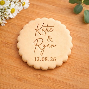 May include: A round biscuit with a decorative, scalloped edge. The biscuit is engraved with the names "Katie & Ryan" and the date "12.08.26". It sits on a wooden surface, with white flowers visible in the background.