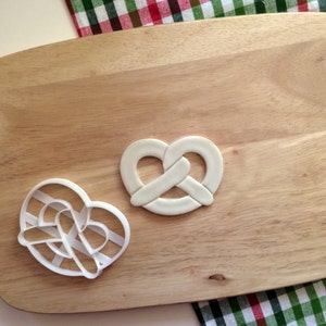 Puede incluir: Dos cortadores de galletas de plástico blanco sobre una tabla de cortar de madera. Un cortador tiene forma de pretzel y el otro tiene forma de corazón.