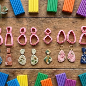 May include: A collection of handmade polymer clay earrings and earring cutters arranged on a wooden surface. The earrings feature various designs, including floral patterns, geometric shapes, and marbled effects. Colorful clay blocks are also present.