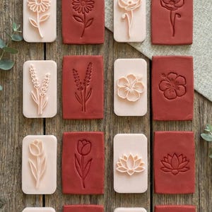 Puede incluir: Colección de sellos rectangulares con motivos florales, incluyendo girasoles y tulipanes. Los sellos son de color rosa claro y marrón rojizo, dispuestos sobre una superficie de madera.