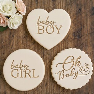 Peut inclure: Trois biscuits clairs pour une baby shower. L'un est en forme de cœur avec "baby BOY", un autre est rond avec "baby GIRL", et le troisième est festonné avec "Oh Baby" et des empreintes de pieds.