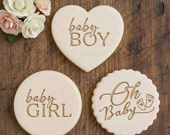 Może przedstawiać: Trzy jasne ciasteczka na baby shower. Jedno jest w kształcie serca z napisem "baby BOY", drugie jest okrągłe z napisem "baby GIRL", a trzecie jest z falbanką z napisem "Oh Baby" i odciskami stóp.