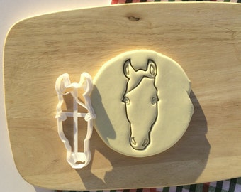 May include: A white plastic horse head cookie cutter on a wooden surface. The cutter is next to a piece of white fondant with a horse head design.
