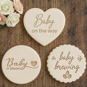 Może przedstawiać: Trzy kremowe ciasteczka z brązowym tekstem. Ciasteczko w kształcie serca z napisem "Baby on the way". Okrągłe ciasteczka z napisami "Baby in bloom" i "a baby is brewing". Bukiet kwiatów znajduje się w lewym górnym rogu.