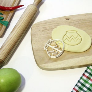 Puede incluir: Un cortador de galletas de plástico blanco con forma de olla con un patrón de zigzag, sobre una tabla de cortar de madera con un rodillo y una manzana verde.